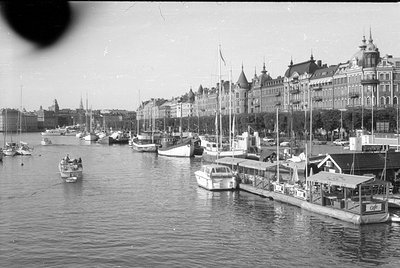 A waterfront scene featuring numerous sailboats and motorboats moored along a quay. In the background, a row of ornate, multi...