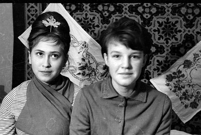 Black and white portrait of two young women posing against a patterned textile backdrop. The woman on the left wears a stripe...