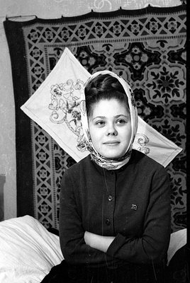 A black and white portrait of a young woman wearing a headscarf and a dark, buttoned cardigan. She is positioned in front of ...