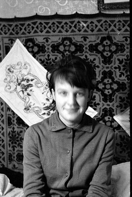 A black and white photograph showing a person lying on a patterned rug and a white embroidered cloth. The image is oriented s...