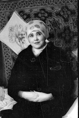 A black and white photograph of a person wearing a patterned headscarf and a dark garment. The subject is positioned diagonal...