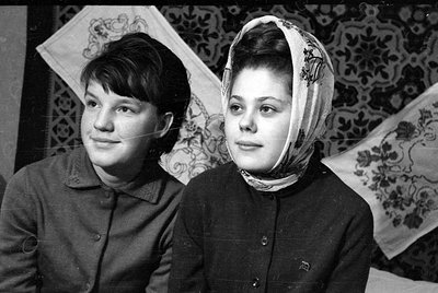 Black and white portrait of two young women. The woman on the right wears a patterned headscarf, and both are dressed in dark...