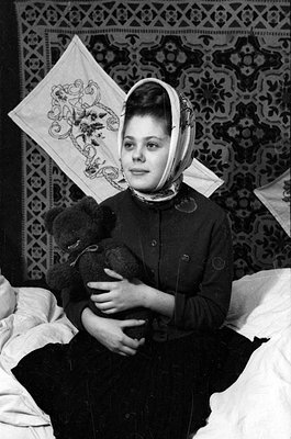 A black and white photograph of a woman wearing a headscarf and a dark cardigan, holding a teddy bear. She is seated against ...