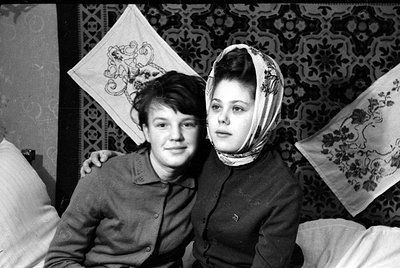 Two young women pose together indoors against a patterned wallpaper. The woman on the right wears a floral headscarf, while t...