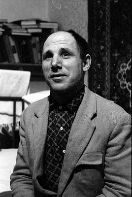 A black and white close-up portrait of a man lying down, looking toward the camera. He is wearing a patterned button-down shi...