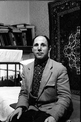 A black and white portrait of a man wearing a light-colored blazer over a dark checkered shirt and bowtie. He is seated in a ...