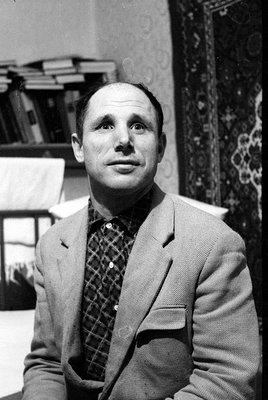 A black and white close-up portrait of a man wearing a light-colored blazer over a dark checkered shirt. He is positioned dia...