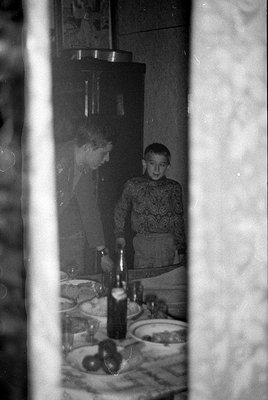 A black and white indoor photograph framed by two light-colored vertical curtains. A woman and a boy in a patterned sweater s...