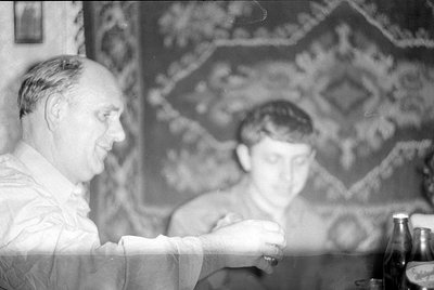 A black and white photograph showing two men indoors. In the foreground, a man in a light-colored shirt is seen in profile, h...