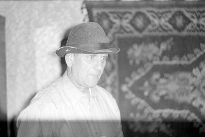 A black and white photograph of a man wearing a fedora and a light-colored button-down shirt. He is positioned in front of a ...