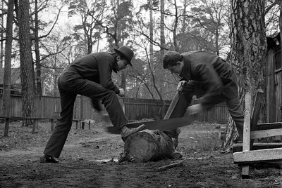 Two men in dark suits and dress shoes use a large two-person crosscut saw to cut through a thick log. One man wears a dark ha...