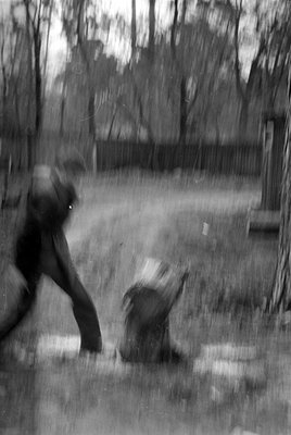 A heavily blurred black and white photograph of a person in motion in a wooded area. A dark fence and vertical tree trunks ar...