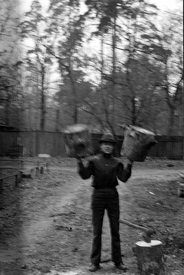 A black and white photograph of a man wearing a dark jacket, trousers, and a brimmed hat. He is standing in a wooded area, ho...