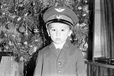 A young boy wearing a peaked cap with a winged emblem and a buttoned suit jacket. He is standing in front of a decorated Chri...