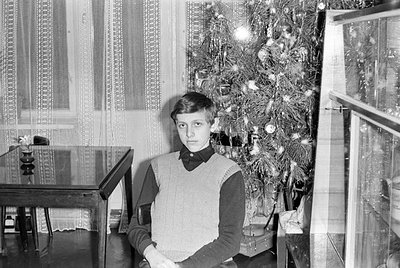 A black and white photograph of a young man wearing a collared shirt and a knit sweater vest. He is seated indoors next to a ...