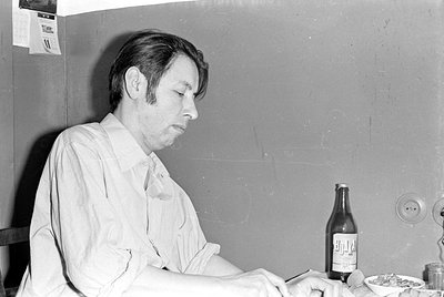 A black and white photograph of a man in a light-colored button-down shirt sitting at a table. On the table is a glass bottle...