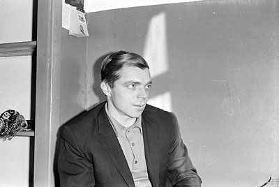 A black and white photograph of a man wearing a dark suit jacket over a light-colored collared shirt. The image is oriented u...