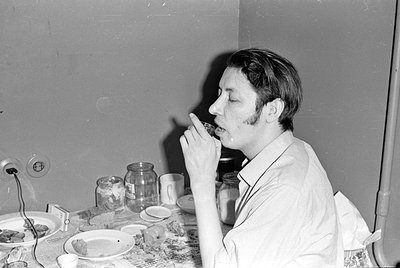 A black and white photograph of a man with sideburns sitting at a table, holding a small glass or cup to his lips. The table ...