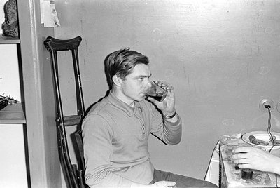 A man in a long-sleeved polo shirt drinks from a glass. He is seated next to a wooden crutch leaning against a wall. A table ...