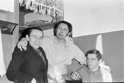 Three men pose together indoors. The man on the left wears glasses and a dark blazer over a patterned shirt; the man in the c...