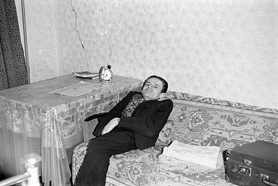 A man in a dark suit and patterned shirt reclines on a patterned sofa. Beside him is a table covered with a lace cloth, holdi...