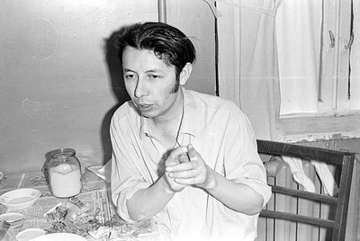 A black and white photograph of a man sitting at a table. He is wearing a light-colored button-down shirt and has his hands c...