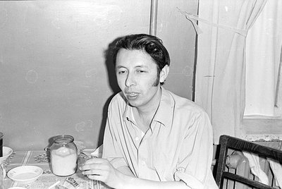 A black and white photograph of a man sitting at a table. He is wearing a light-colored button-down shirt. On the table are a...