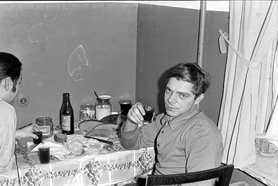 A black and white photograph of two men sitting at a table covered with a patterned cloth. On the table are a bottle, several...