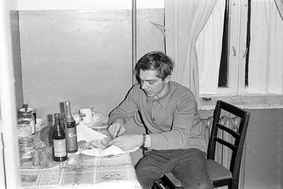 A man in a long-sleeved shirt sits at a table covered with a patterned cloth. On the table are two bottles, glasses, a cup an...