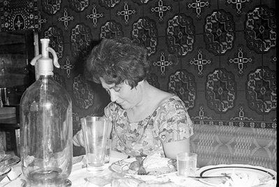 A woman in a floral print dress sits at a table covered with a white cloth. In the foreground stands a large glass seltzer bo...