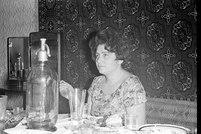 A woman in a floral print dress sits at a table in front of a dark, patterned textile wall hanging. A large glass seltzer bot...