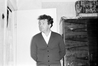 A black and white photograph of a man with curly hair wearing a dark cardigan over a white collared shirt. He is standing ind...