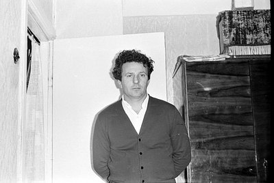 A black and white portrait of a man with curly hair wearing a dark suit jacket and a white collared shirt. He is positioned a...