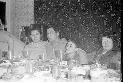 A black and white photograph, oriented upside down, showing three people seated at a table. The background consists of a dark...