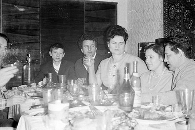 A black and white photograph of five people seated around a dining table. A man in the center holds up a small glass, while b...