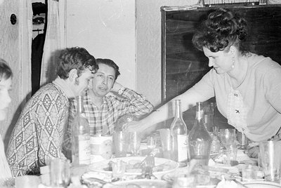A black and white indoor photograph of three people seated at a table with bottles and glasses. A woman on the right reaches ...