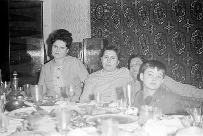 A black and white photograph of four people seated at a dining table. Two women and two males are visible against a backdrop ...