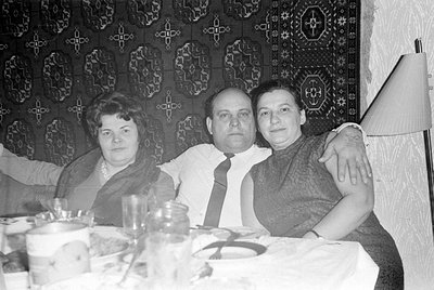 Three adults sit at a table covered with a white cloth and glassware. A man in a white shirt and tie has his arm around a wom...