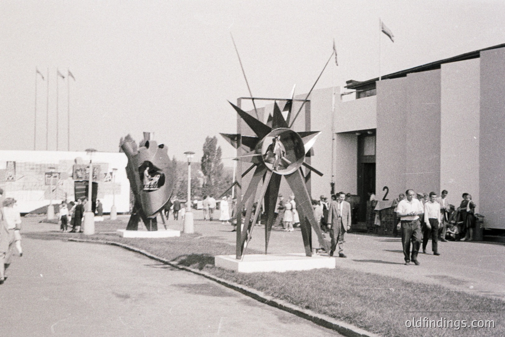 Two tall abstract sculptures standing on a paved forecourt, a low modernist building with vertical striped cladding and a row of flagpoles behind them