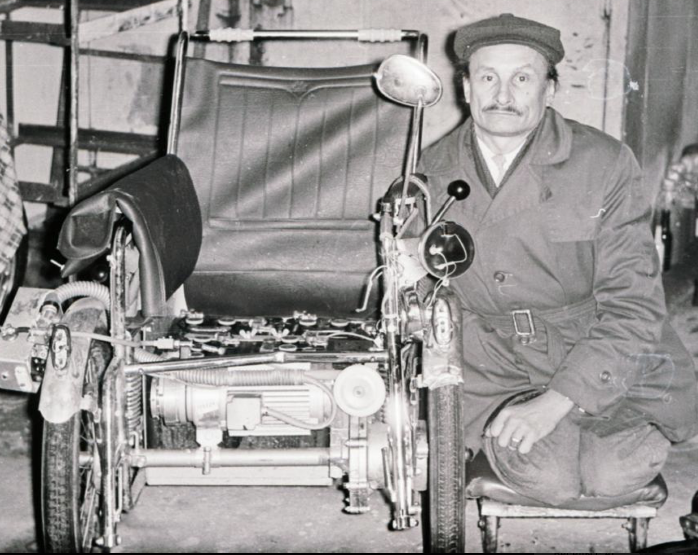 A man kneeling on a low wheeled board beside a partly dismantled wheelchair in a workshop, its motor and contactors exposed