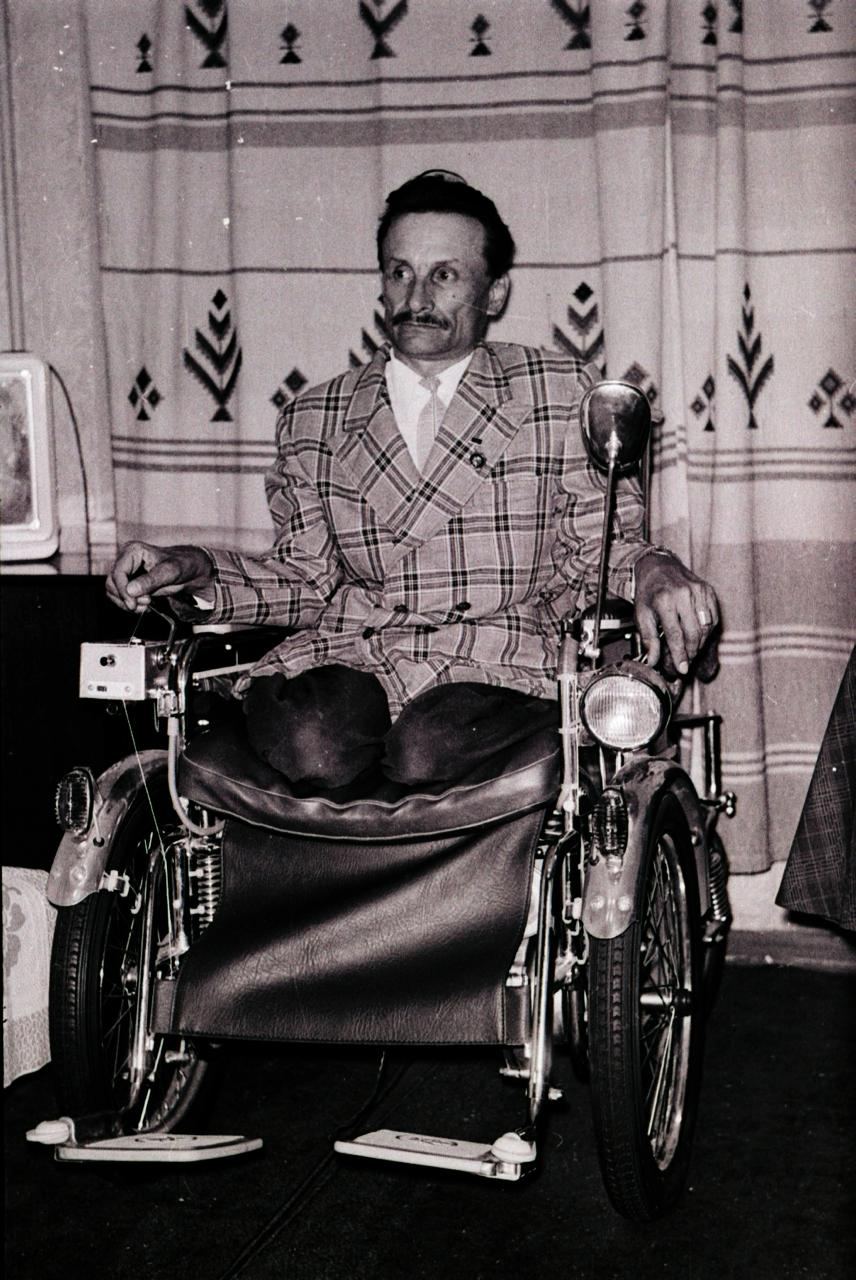 The same man in a double-breasted checked jacket and tie, seated in the same wheelchair, now fully assembled with fenders, headlamp and mirror, in front of a patterned curtain