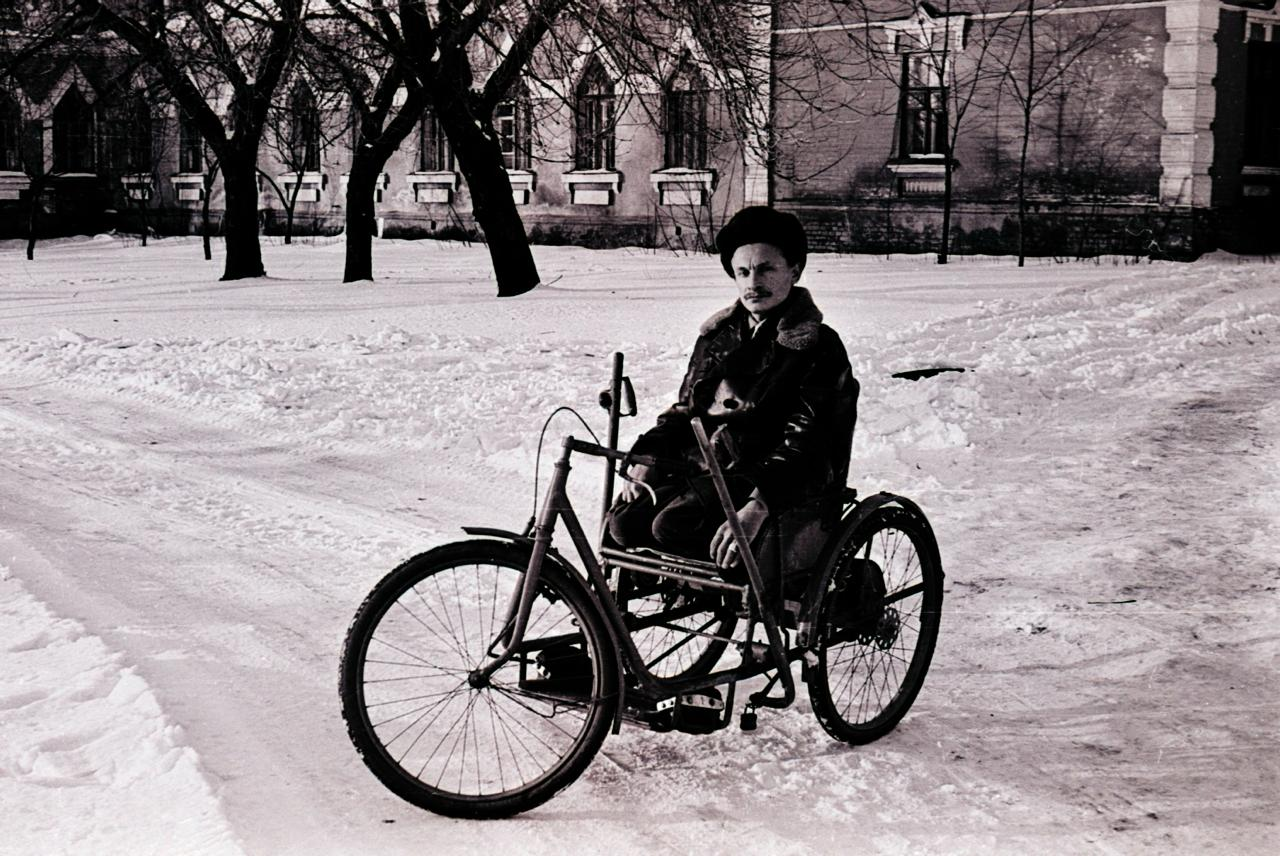 A man on a lever-driven tricycle wheelchair on a snow-covered path, in front of a long red-brick building with pointed-arch windows and bare trees