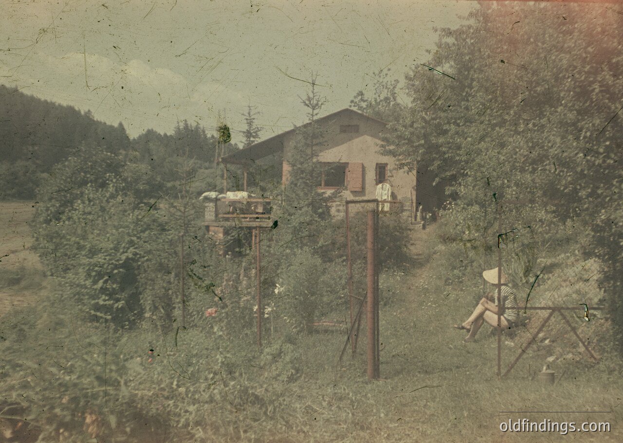 Vintage sepia-toned photo of a rural homestead with a two-story wooden house and elevated wooden platform. Early 20th-century farm equipment, including a spinning wheel and wooden cart, suggests agricultural life. Lush greenery and dense trees frame the scene, likely Eastern European countryside.