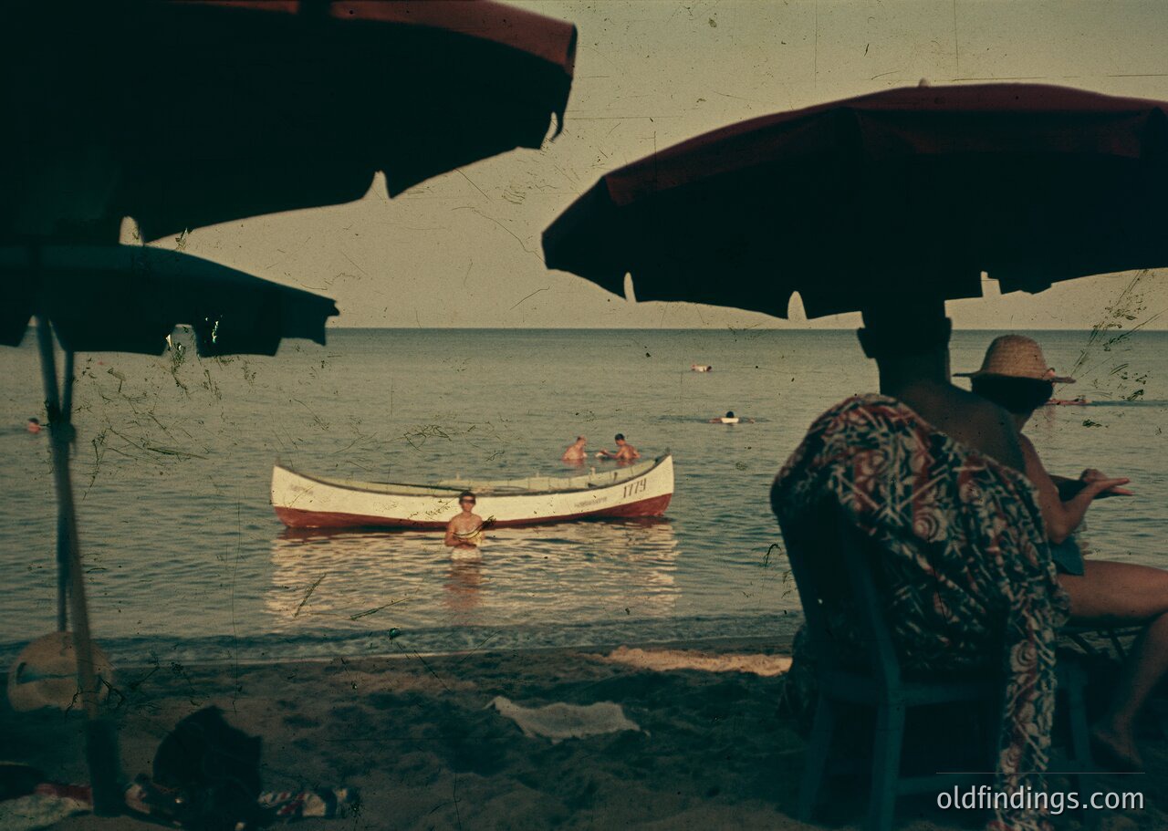Vintage seaside scene with two men by the water’s edge under striped umbrellas. One man stands in shallow water near a small wooden boat; the other sits on a rock, gesturing. Mid-20th century beach attire and boat design suggest coastal leisure.