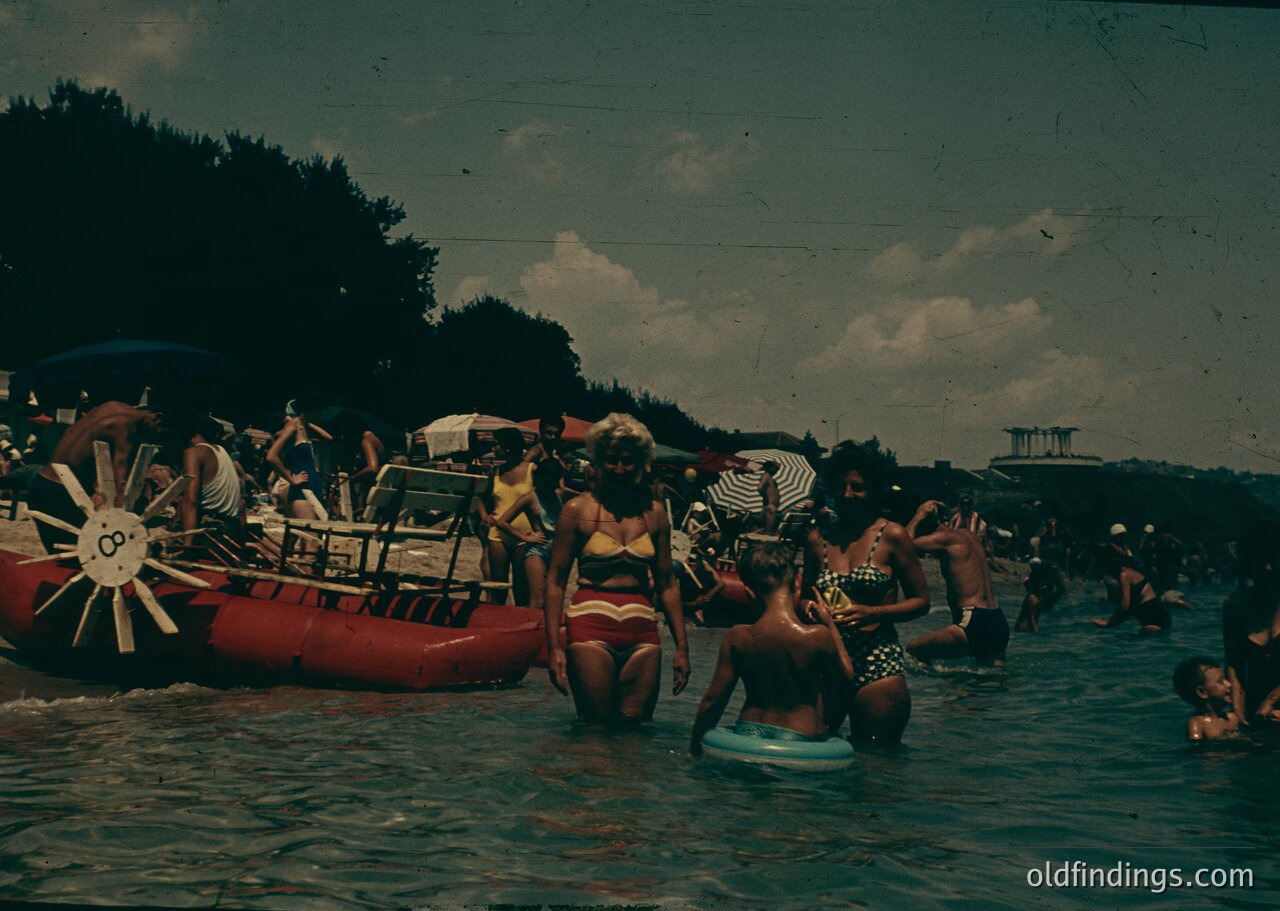 Vintage beach scene featuring a red inflatable raft with a sunburst logo, mid-20th century swimwear, and a lively crowd enjoying shallow waters. Trees and beach umbrellas suggest a resort atmosphere, likely 1960s–1970s.