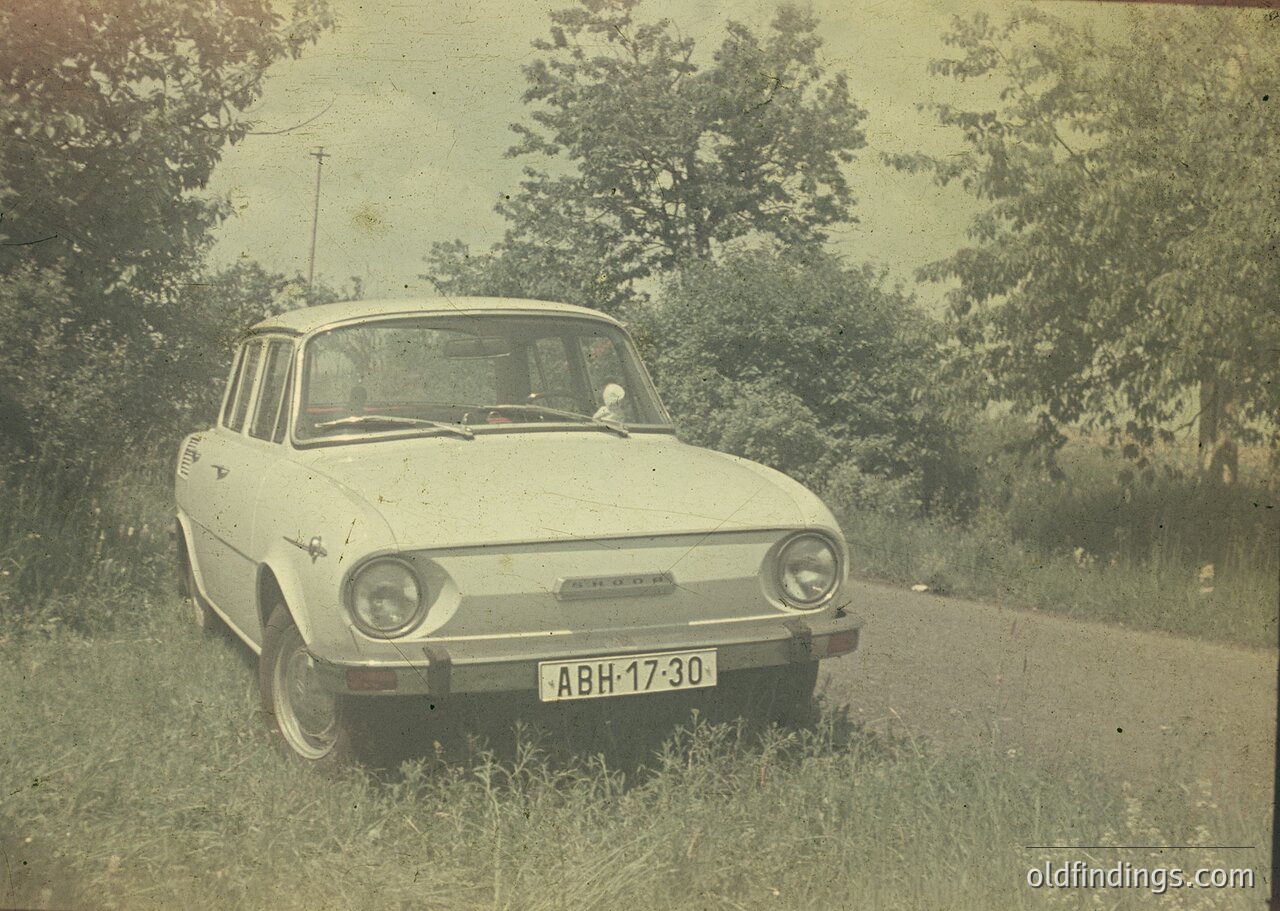 1970s-era Zastava 750 sedan parked on a rural road, identifiable by round headlights and "Zastava" badge. Rustic surroundings suggest Eastern European countryside. Vintage sepia tone enhances nostalgic appeal.