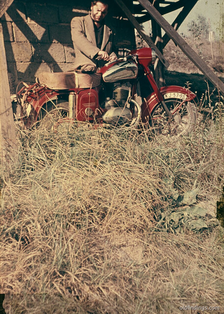 Mid-century red motorcycle with classic step-through frame, parked in dry grass beside a stone wall. Man in dark suit and cap poses beside it, suggesting a rural or countryside setting, likely 1950s–1960s.