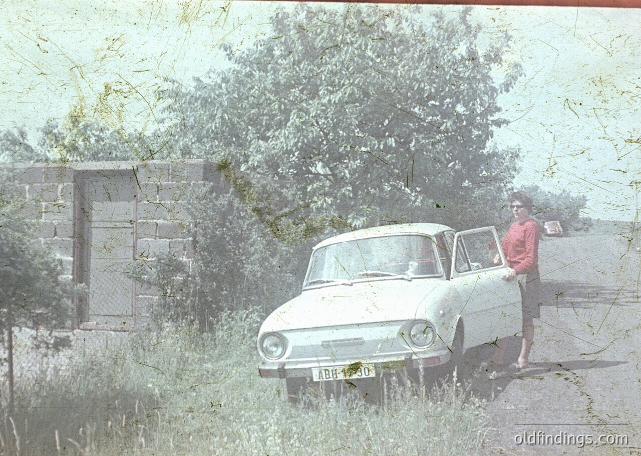 Vintage 1960s-era sedan parked beside a concrete wall and overgrown vegetation. Man in retro sunglasses and striped shirt poses near the car’s front fender. Handwritten license plate visible. Sepia-toned photo suggests archival or nostalgic appeal.