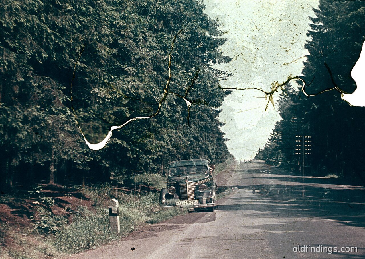 Vintage sepia-toned photo of a mid-20th-century roadside scene, featuring a classic sedan parked beside a mailbox. Dense evergreen trees frame both sides of the narrow, winding road, with a faintly visible signpost ahead. The aged film grain and sepia tone suggest or American countryside.