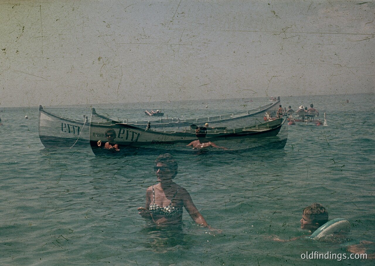 Vintage seaside scene featuring two wooden boats labeled "ПИТТ" in Cyrillic, likely Bulgarian. A woman in a patterned swimsuit swims near the foreground, while others relax or swim in the water. Distressed film grain and faded tones suggest mid-20th century, possibly . Coastal leisure activity with a nostalgic, retro aesthetic.